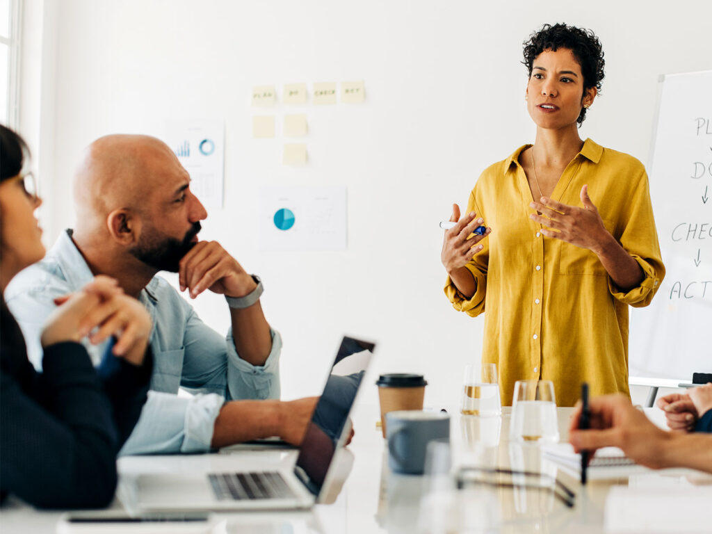 A female facilitator in a yellow blouse leads a corporate workshop, presenting to a diverse team with laptops and notes.