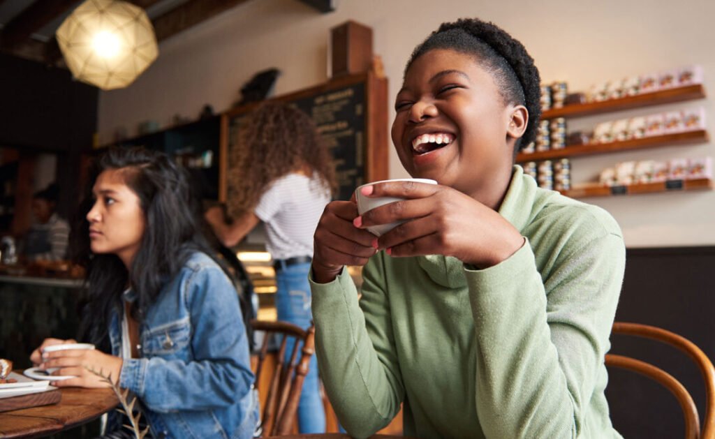 A woman in a green sweater laughing with coffee, representing customer-centric leadership in the food and beverage industry.